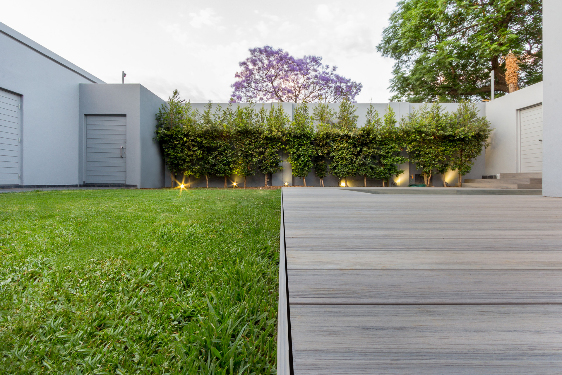 Small backyard with bushes against a tall fence with a composite decking lining a grass area. 