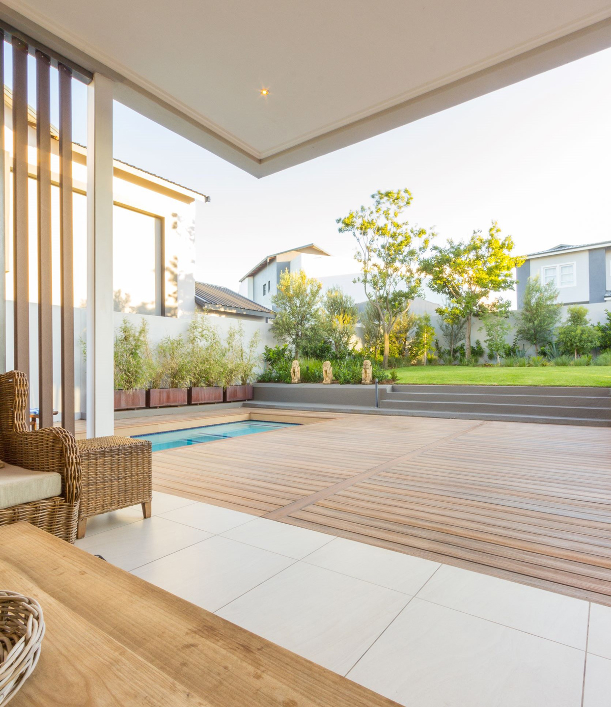 Outdoor living space with light colored composite decking adjacent to a pool with green trees in the background.