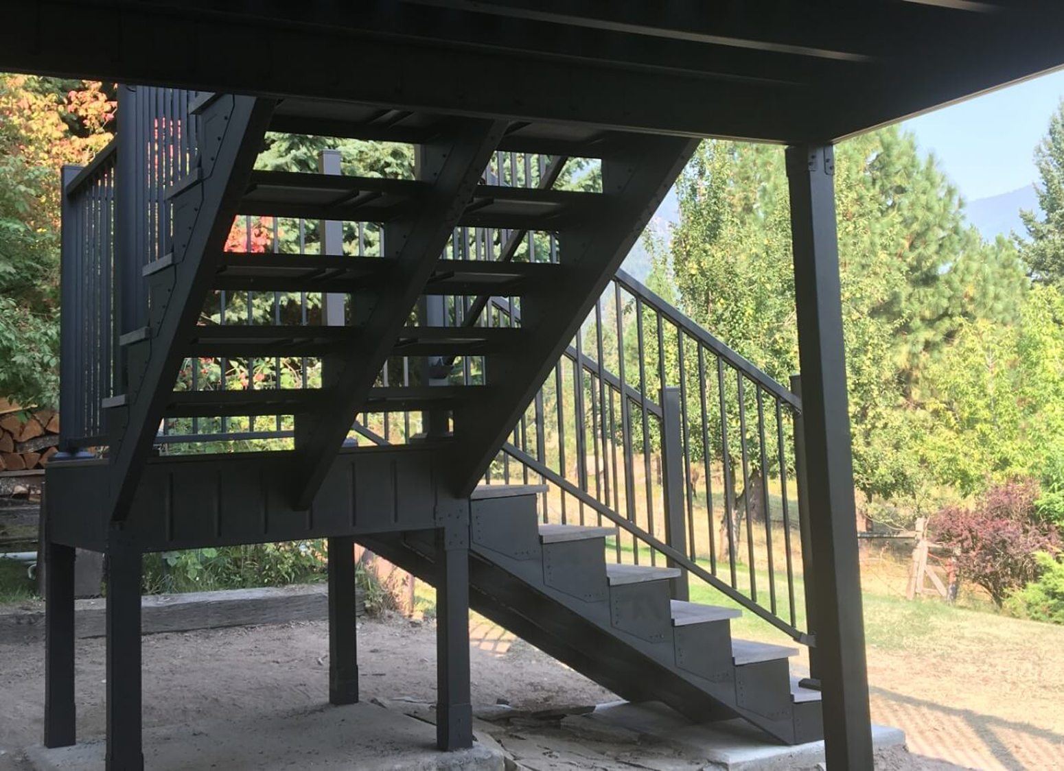 View from beneath deck stairs showing black steel deck framing with trees in the background.