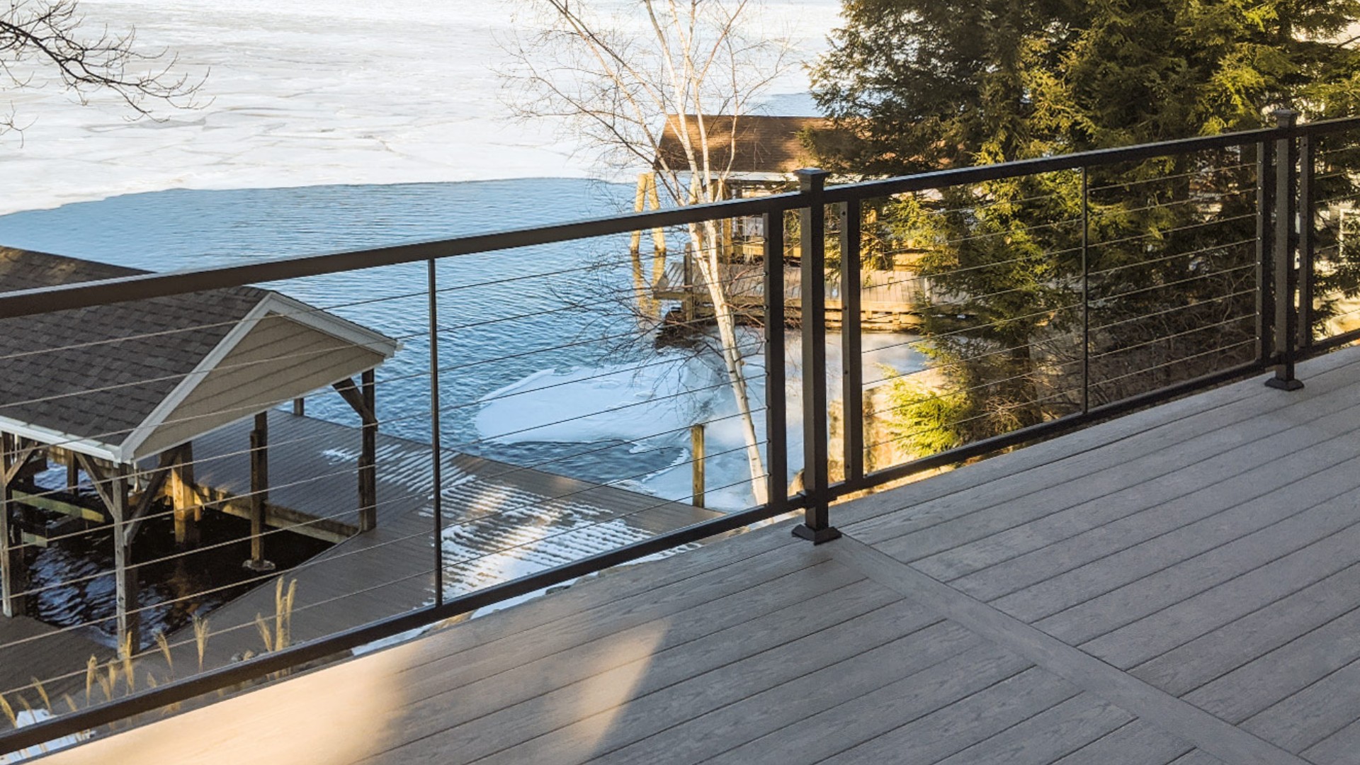 Balcony with sleek cable railing overlooking a lake with trees and a boathouse in the background.