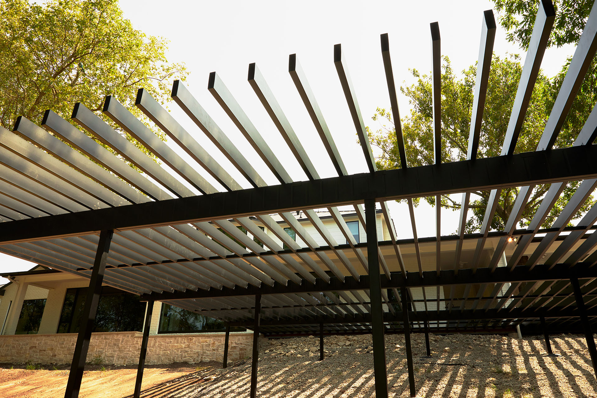 View from beneath a large steel pergola with horizontal beams casting shadows on the ground.